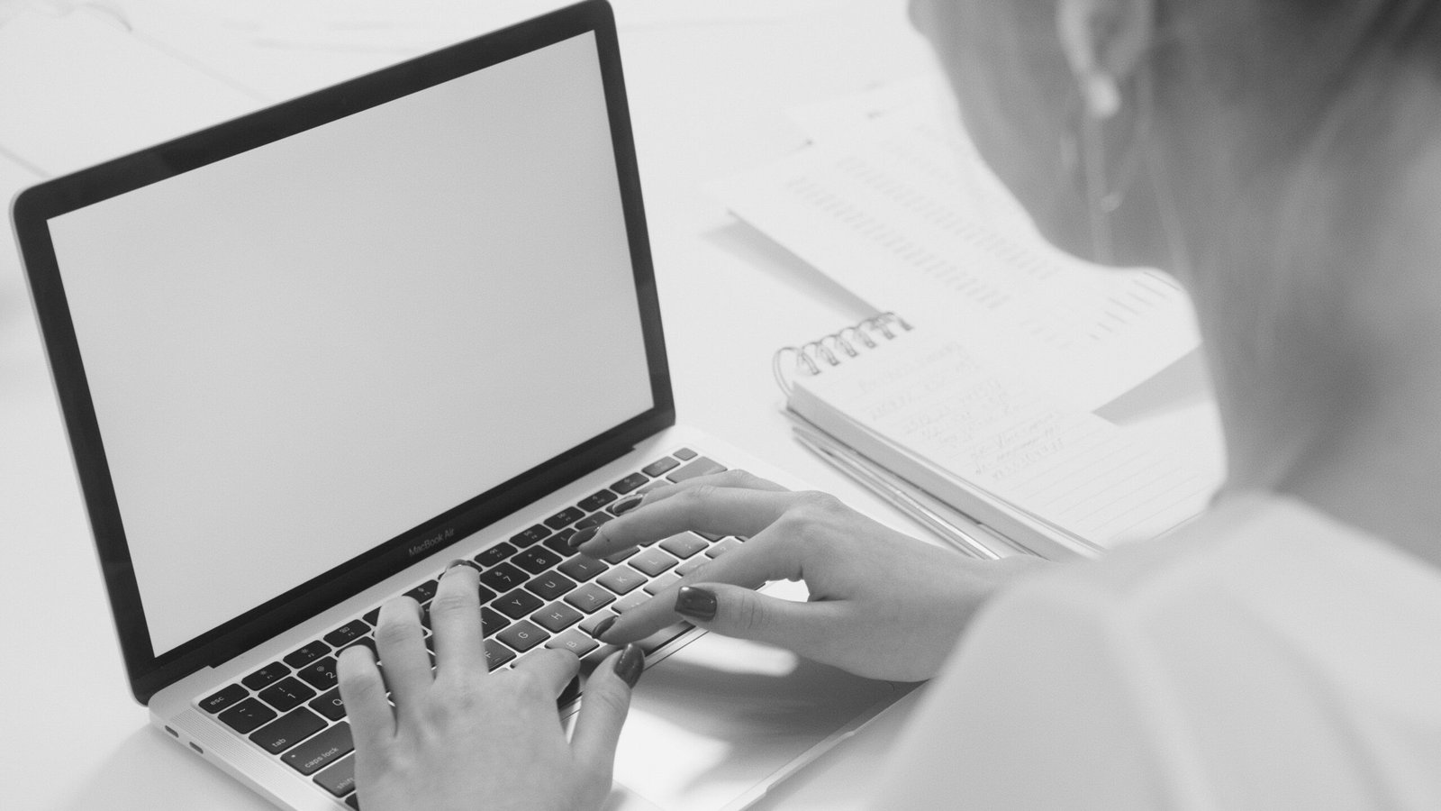 Monochrome image of a person typing on a laptop with a notebook nearby.